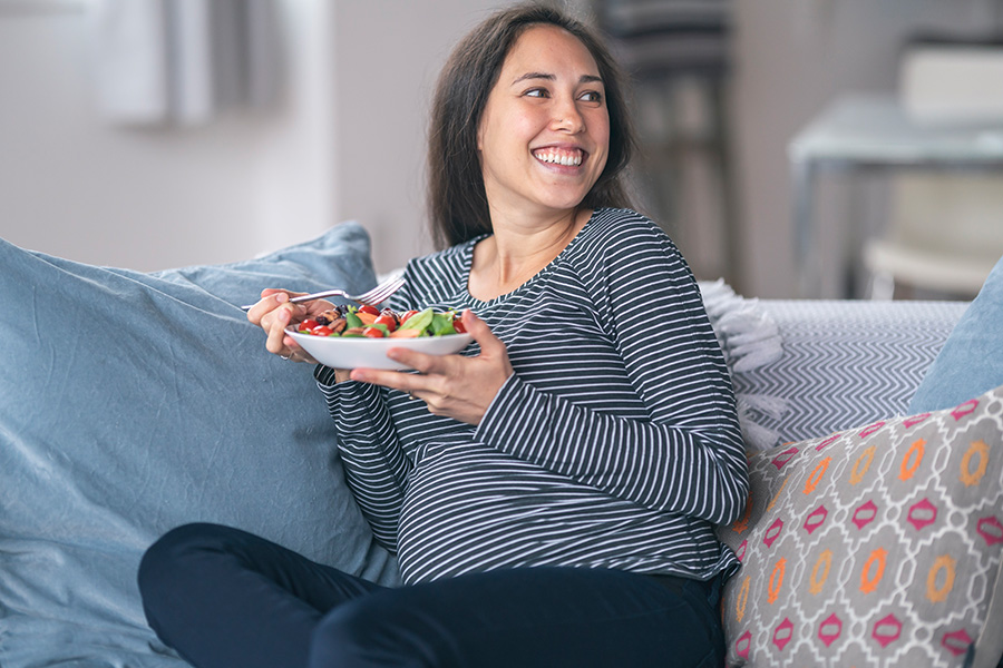 Schwangere Frau sitzt auf Couch und isst einen Salat